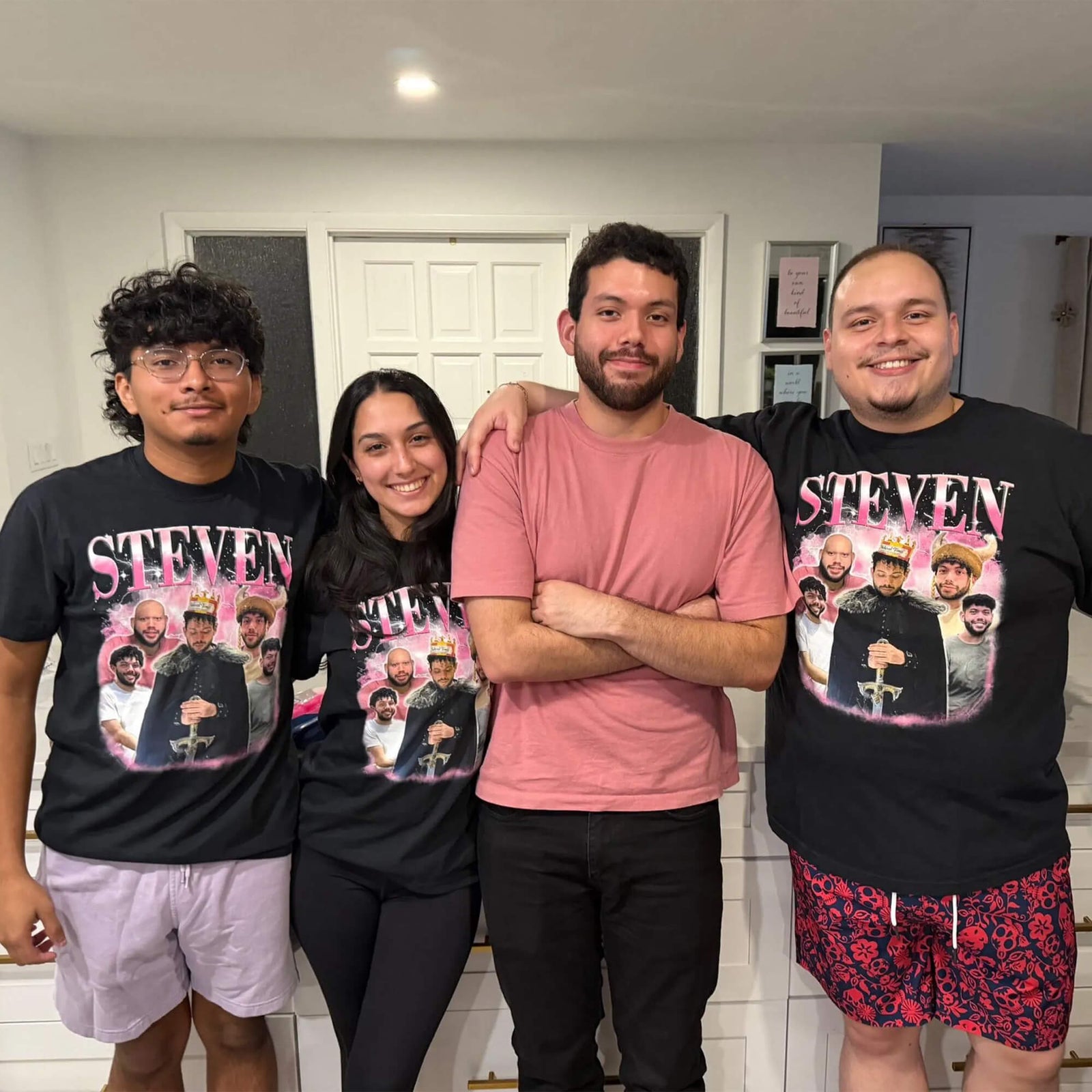 Four men standing together indoors wearing matching black graphic T-shirts with a large 'STEVEN' photo print on the front. The shirts feature a portrait of a man in a black robe with several children around him, with pink and white text.