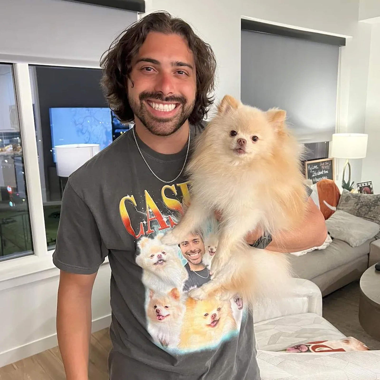 A smiling man holding a fluffy cream Pomeranian dog in a modern living room. The man is wearing a dark gray graphic t-shirt featuring a custom photo print with the text 'CASE' and images of dogs.