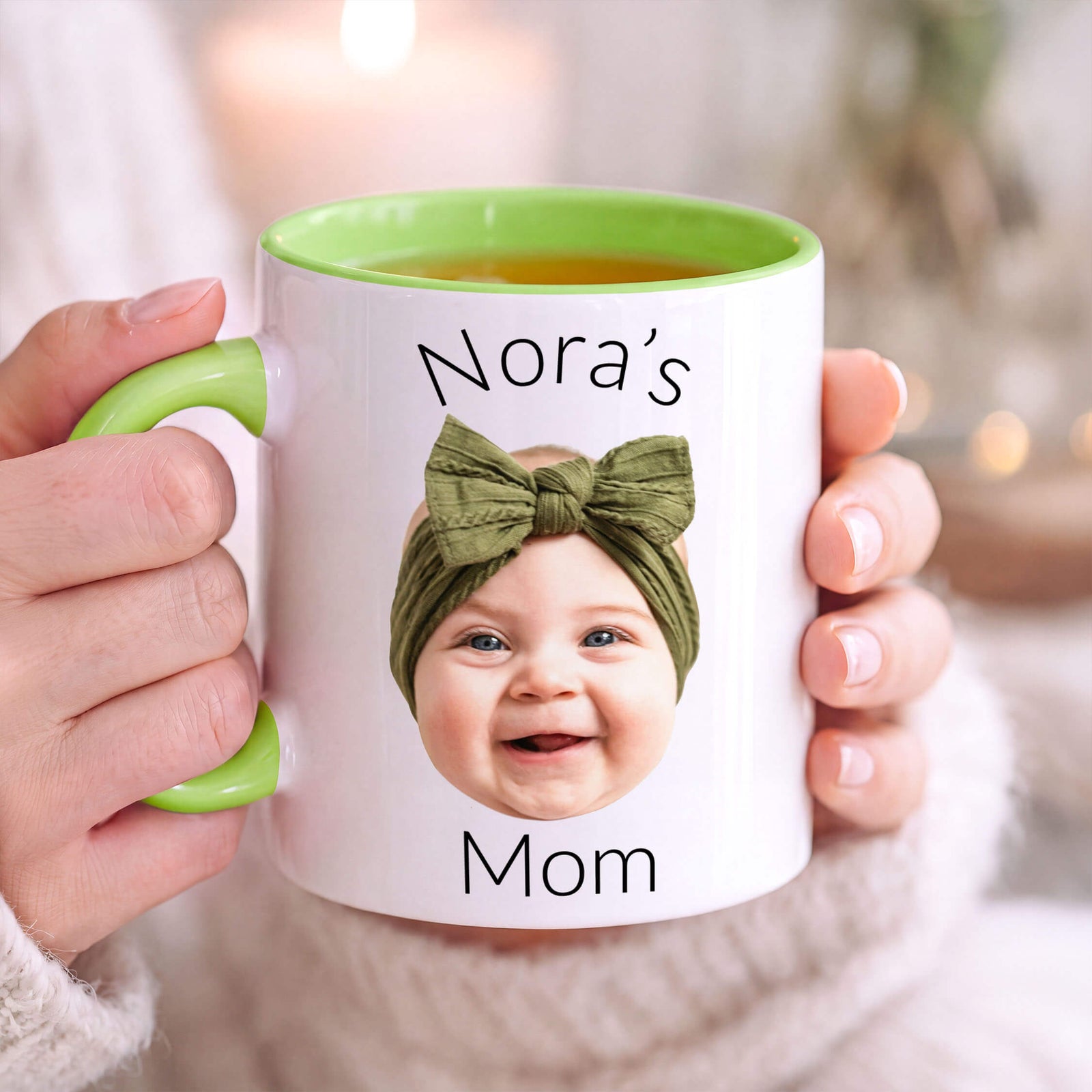 A person holds a white ceramic mug with a green handle and green interior. The mug is printed with the text 'Nora's Mom' and features a photo of a smiling baby wearing an olive green bow headband.