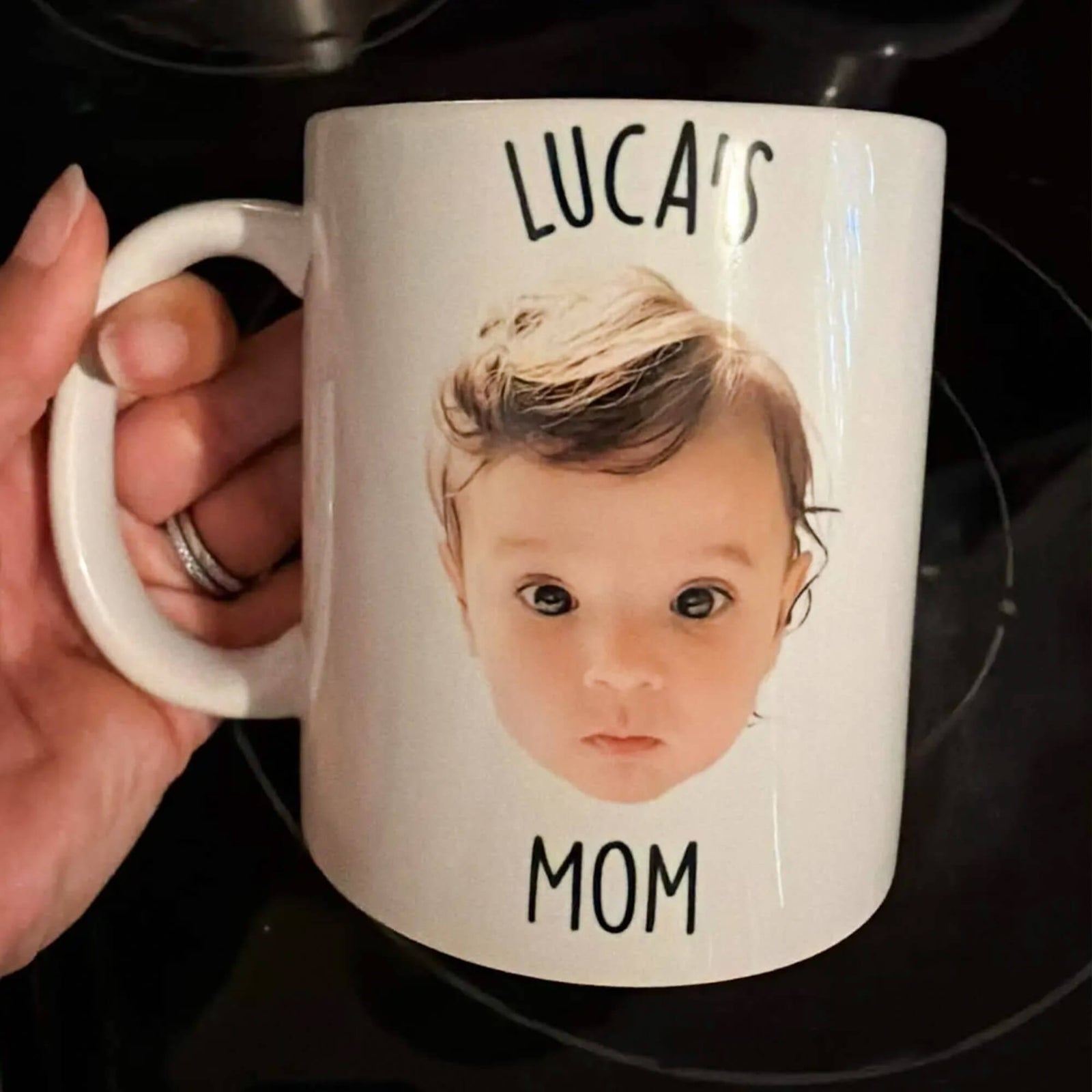A hand holds a white ceramic mug printed with a baby photo and the name in black text, set against a dark stovetop background.
