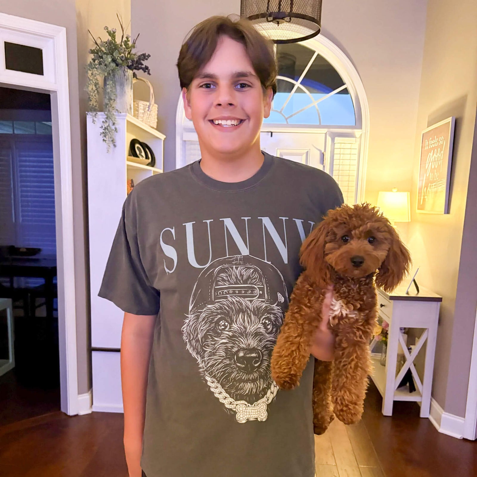 Boy wearing a charcoal pet-portrait T-shirt with a dog illustration while holding a small curly brown dog at home.