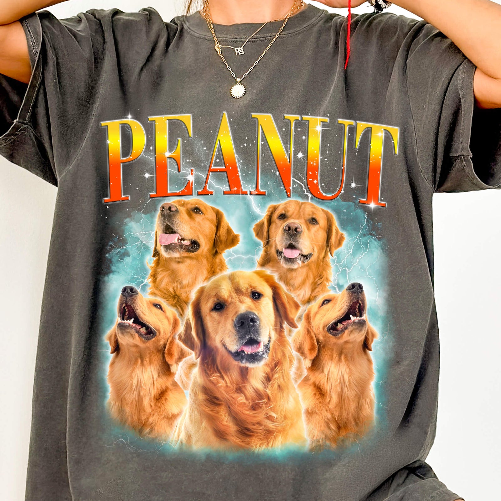 Close-up of a charcoal T-shirt printed with PEANUT in orange-to-yellow lettering above a collage of five golden retrievers on a glowing turquoise lightning background.