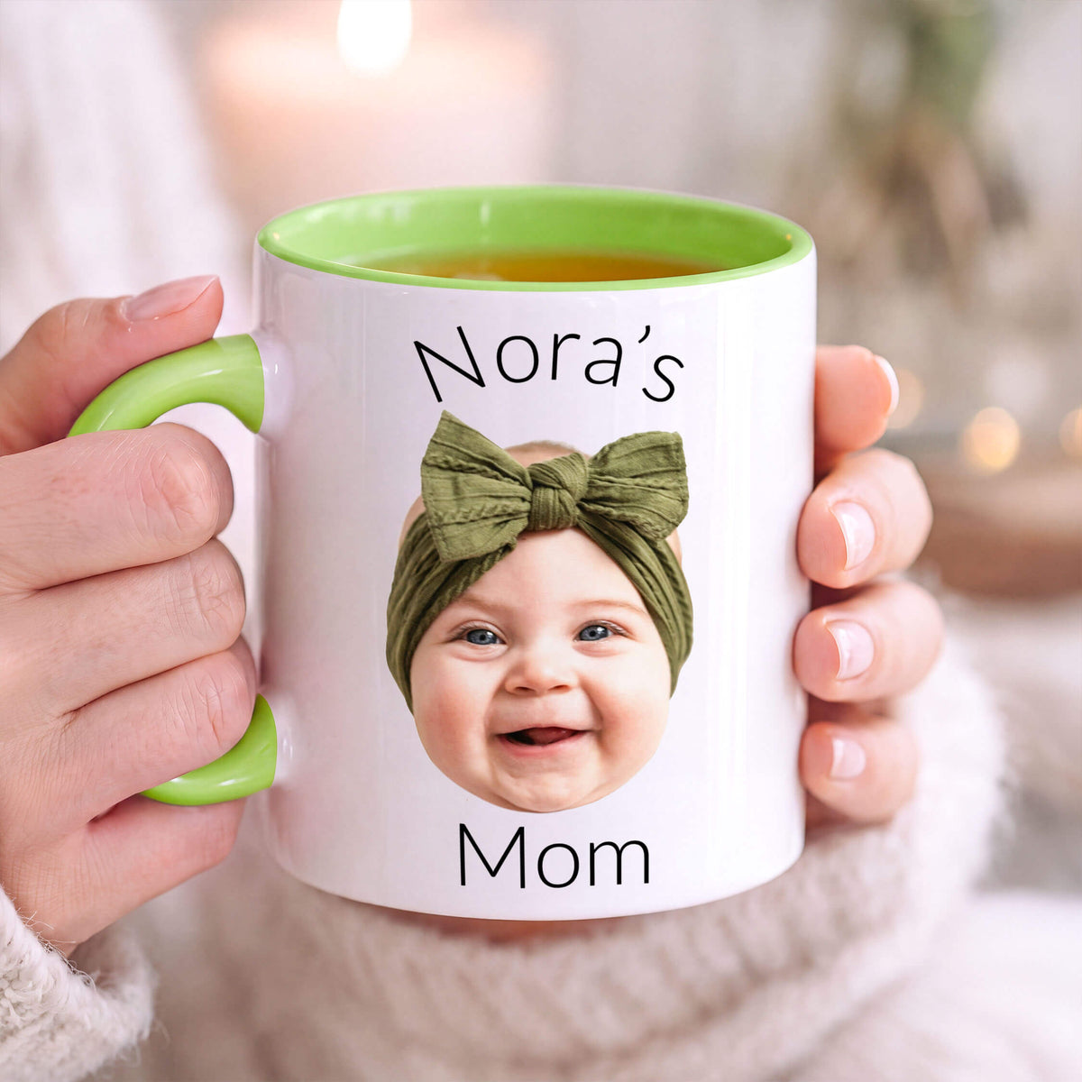 A person holds a white ceramic mug with a green handle and green interior. The mug is printed with the text 'Nora's Mom' and features a photo of a smiling baby wearing an olive green bow headband.