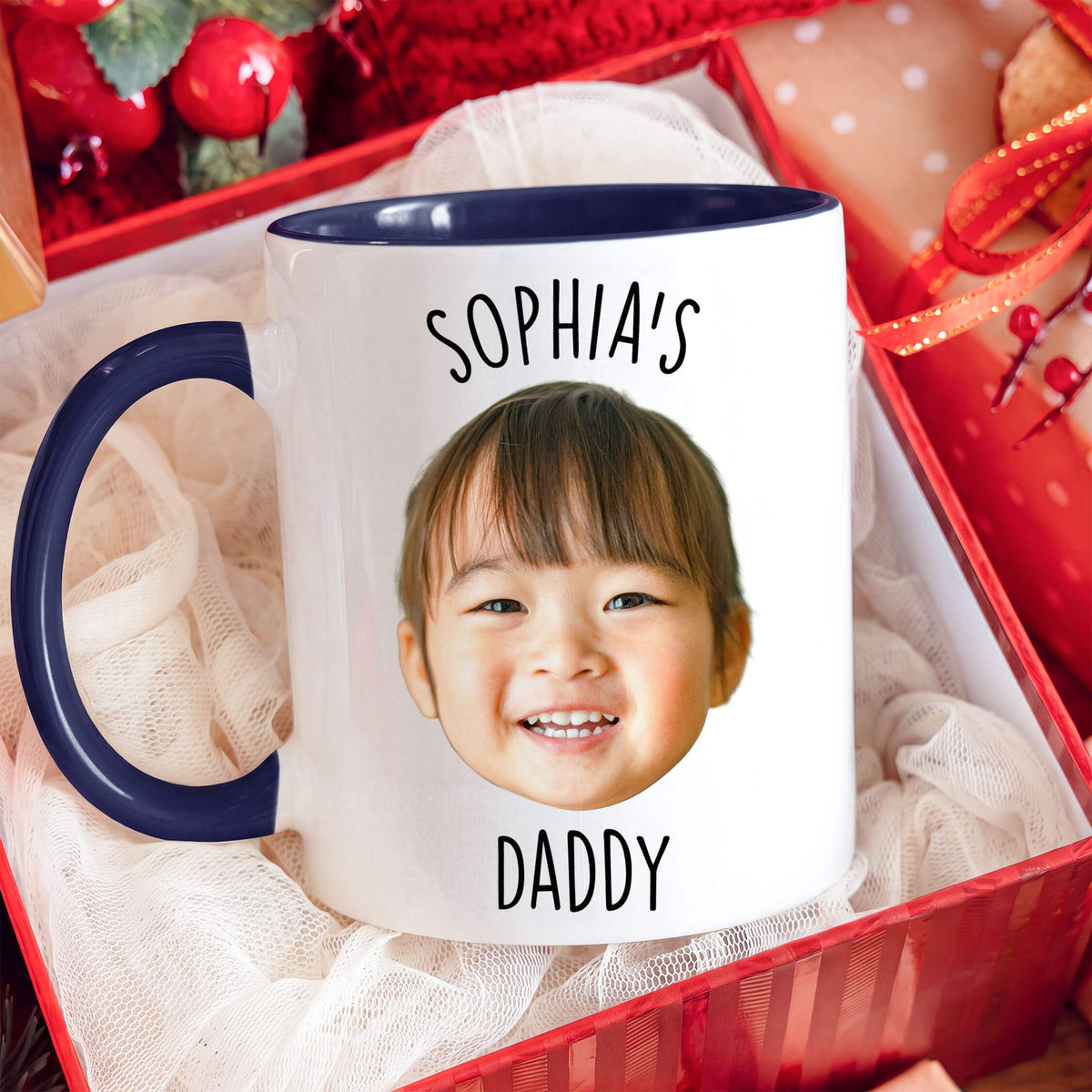 A white ceramic mug with a navy blue handle and interior, printed with the text 'SOPHIA'S DADDY' and a smiling child's face. The mug sits in a red gift box filled with white tulle fabric.