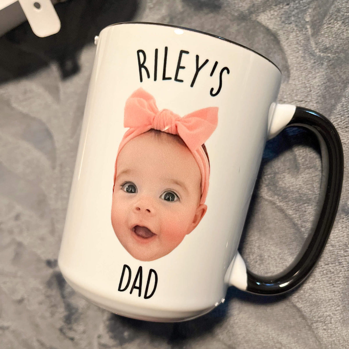 A white ceramic mug with a black handle and black rim, printed with the text 'RILEY'S DAD' and a baby photo wearing a pink bow headband, resting on a gray textured surface.