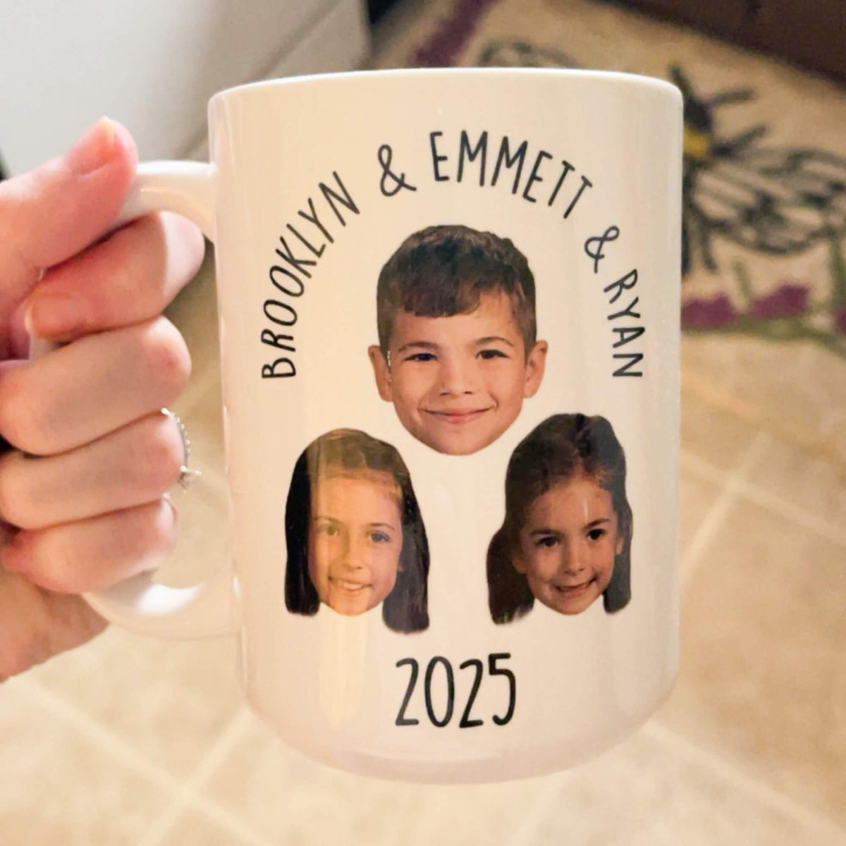 A hand holds a white ceramic mug with custom printed photos of three children and the names around the top, with '2025' printed at the bottom.