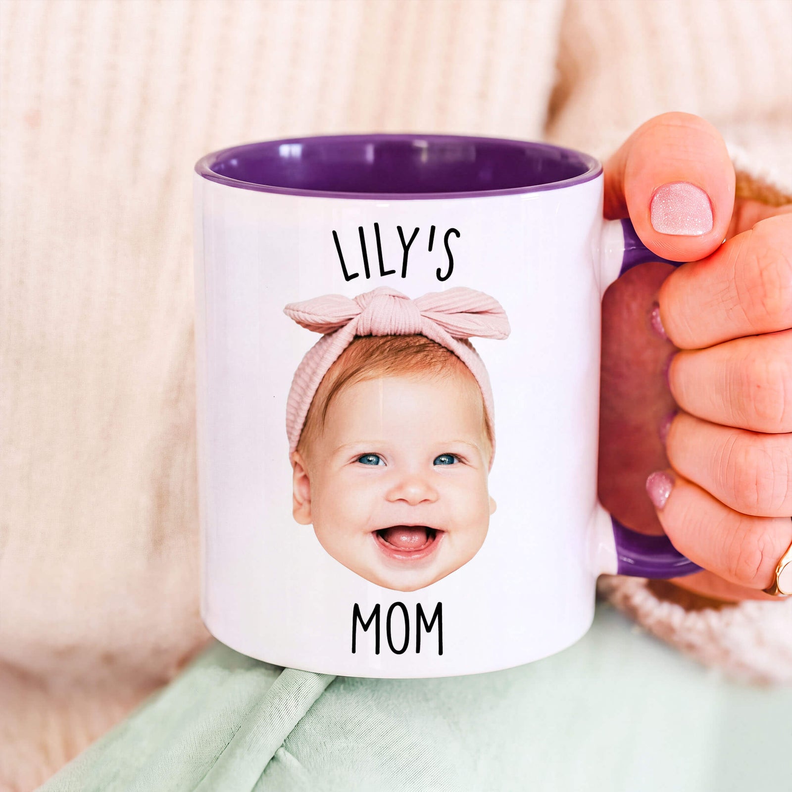 A white ceramic mug with a purple interior and handle, printed with the text 'LILY'S MOM' and a photo of a smiling baby wearing a pink bow headband. A hand holds the mug against a soft beige knit background.